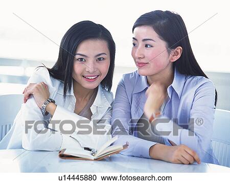Stock Image - Image of Two Young Adult Women Sitting In Front of a Calendar, One Looking at Camera, Smiling, Front View. Fotosearch