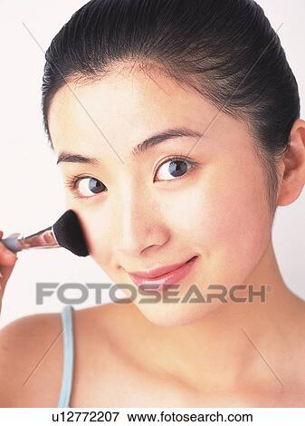 Stock Photo - Japanese woman applying blusher on cheek, looking at camera. Fotosearch