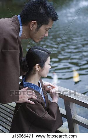 Stock Image - Mid Adult Couple in Yukata, Looking at Ornamental Crap in Pond, Side View. Fotosearch
