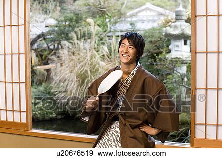Stock Photo - Man in Yukata holding a Japanese fan and sitting at the window, front view, Japan. Fotosearch