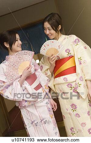 Stock Image - Two women in Yukata standing side by side and holding Japanese folding fans, front view, Japan. Fotosearch