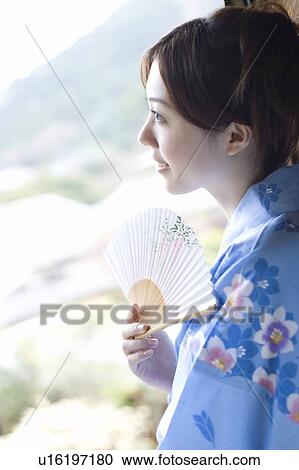 Stock Image - Woman in Yukata holding a Japanese folding fan and looking outside from the window, side view, Japan. Fotosearch
