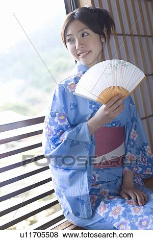 Stock Photo - Woman in Yukata holding a Japanese folding fan and sitting at the window, front view, Japan. Fotosearch