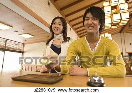 Stock Photo - Young couple checking into a hotel, man holding a card, front view, Japan. Fotosearch