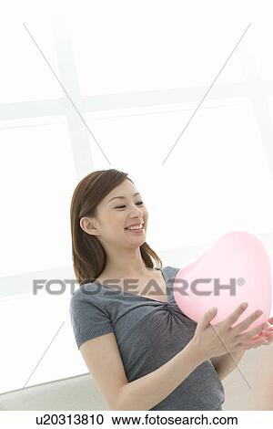 Stock Image - A woman holding a pink heart, smiling, front view. Fotosearch
