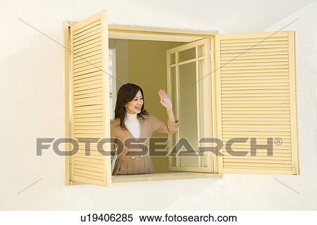 Stock Photography - A woman smiling and waving the hand at the window, front view. Fotosearch