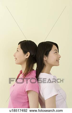 Stock Image - Portrait of Two Young Twin Sisters Back to Back, Smiling, Side View. Fotosearch