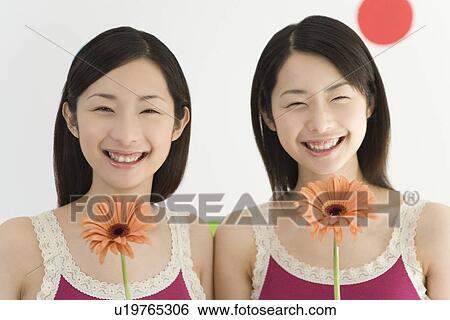 Stock Photograph - Portrait of Two Young Twin Sisters Holding Flowers of Gerbera, Smiling, Front View. Fotosearch