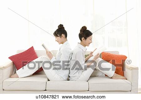 Twin sisters sitting on couch and reading books, back to back, smiling, side view View Large Photo Image Stock Image - Twin sisters sitting on couch and reading books, back to back, smiling, side view. Fotosearch