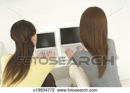 Stock Image - Two women sitting on a couch and using laptops, rear view, white background, high angle view. Fotosearch