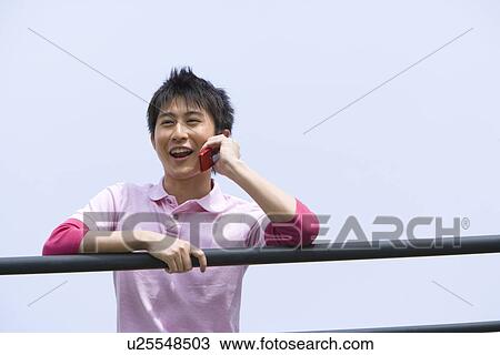 Stock Image - Man leaning on the railing and using a mobile phone under the blue sky, front view, blue background, Japan. Fotosearch