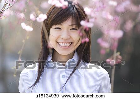 Stock Photo - Portrait of a woman standing under the cherry tree, smiling and looking at camera, front view, Japan. Fotosearch