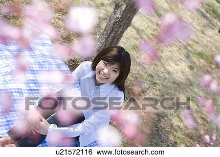Stock Photograph - Portrait of a young woman sitting on a sheet under the cherry tree, smiling and looking at camera, high angle view, differential focus, Japan. Fotosearch