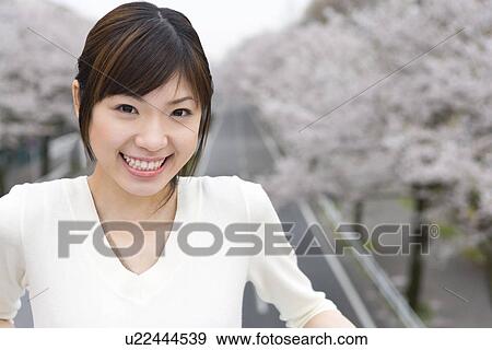 Stock Photo - Portrait of a young woman smiling and looking at camera, road lined with cherry trees in background, front view, Japan. Fotosearch