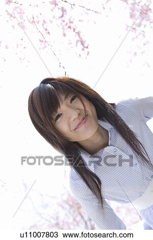 Stock Image - Portrait of a young woman under the cherry tree, smiling and looking at camera, low angle view, white background, Japan. Fotosearch