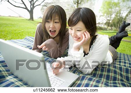 Stock Image - Two young women looking at a laptop, lying on sheet, front view, Japan. Fotosearch