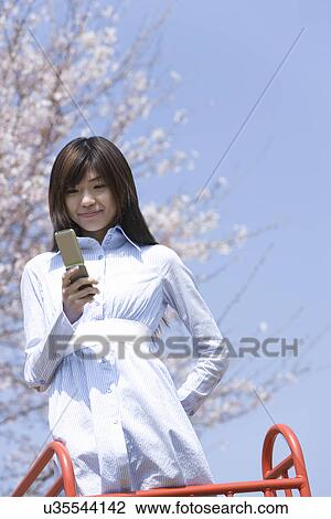 Stock Image - Woman smiling and using a mobile phone under the cherry tree, low angle view, blue background, Japan. Fotosearch