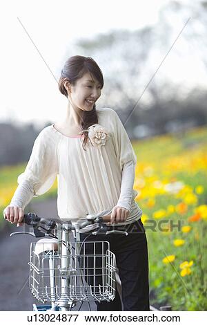 Stock Photo - Woman with bicycle smiling and looking at field of corn poppy, front view, Japan. Fotosearch