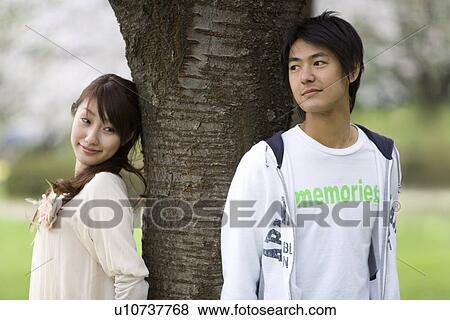 Stock Photo - Young couple leaning on a tree trunk, front view, side view, Japan. Fotosearch