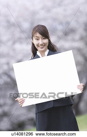 Stock Photograph - Portrait of a woman holding a blank board in the field of cherry blossoms, smiling and looking at camera, front view, Japan. Fotosearch