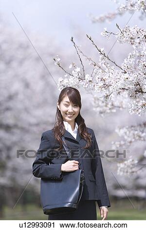 Stock Image - Portrait of a woman standing in the field of cherry blossoms, smiling and looking at camera, front view, Japan. Fotosearch