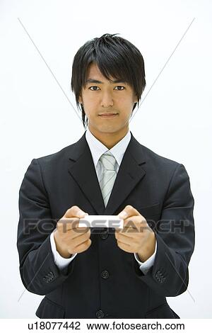 Stock Image - Portrait of a young businessman holding a business card, looking at camera, front view, white background. Fotosearch
