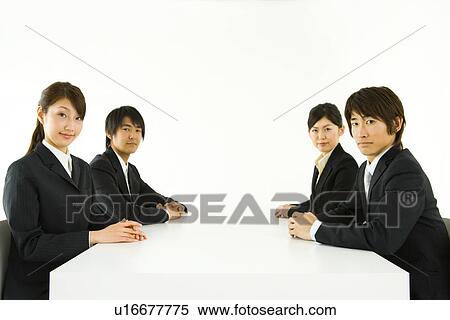 Stock Photography - Portrait of four young business people sitting at table, looking at camera, side view, white background, Japan. Fotosearch