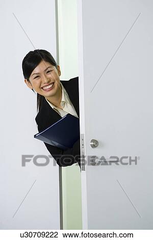 Stock Image - Portrait of young businesswoman behind the door, smiling and looking at camera, front view, Japan. Fotosearch