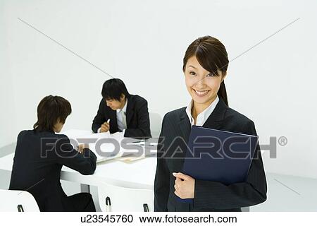 Stock Image - Three young business people in meeting, woman looking at camera, front view, rear view, white background, Japan. Fotosearch