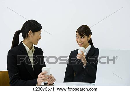 Stock Image - Two young businesswomen sitting at desk, smiling and holding paper cups, front view, side view, white background, Japan. Fotosearch