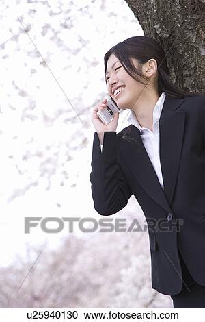 Stock Image - Woman leaning on trunk of a cherry tree and using mobile phone, side view, Japan. Fotosearch