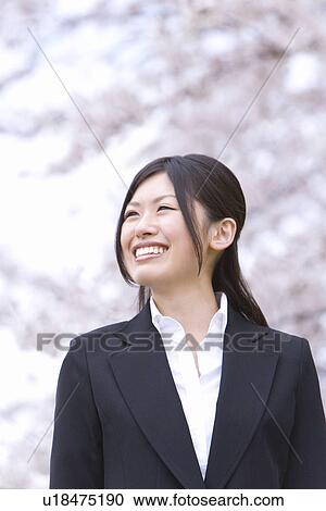 Stock Image - Woman smiling and standing under the cherry tree, looking away, front view, Japan. Fotosearch