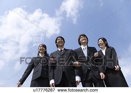 Stock Photo - Young business people standing side by side under the blue sky, arm in arm, low angle view, Japan. Fotosearch