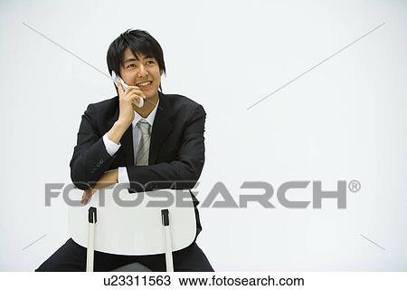 Stock Image - Young businessman sitting on chair and using mobile phone, front view, white background, copy space. Fotosearch