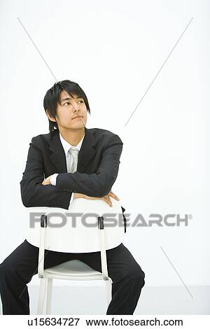 Stock Photo - Young businessman sitting on chair, looking away, front view, white background, copy space. Fotosearch