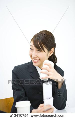 Picture - Young businesswoman sitting at desk, holding paper cup and mobile phone, front view, white background, Japan. Fotosearch