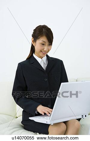 Stock Photography - Young businesswoman sitting on couch and using laptop, front view, white background, Japan. Fotosearch