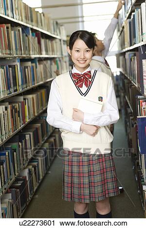 Stock Image - Portrait of a teenage girl holding a book in library, smiling and looking at camera, boy in background, front view, side view. Fotosearch