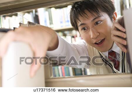 Stock Photograph - Teenage boy holding a book in bookcase, front view. Fotosearch
