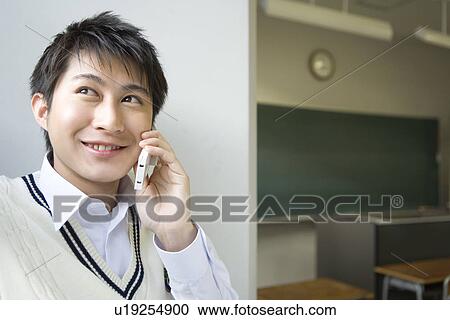 Stock Image - Teenage boy using mobile phone in classroom, front view. Fotosearch