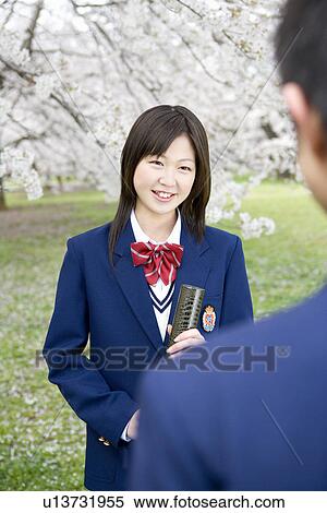 Stock Photography - Teenage girl and boy standing under cherry trees, girl holding a diploma, front view, rear view, Japan. Fotosearch