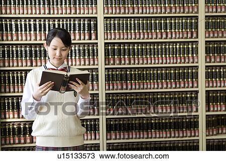 Stock Image - Teenage girl reading a book in front of bookcase, front view. Fotosearch