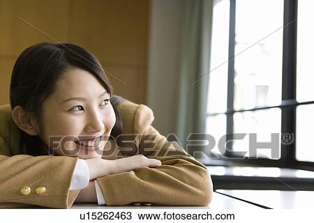Stock Image - Teenage girl sitting at desk and leaning on arm in classroom, smiling and looking away, front view. Fotosearch