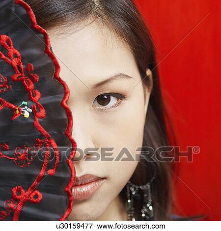 Stock Photo - Portrait of a young woman covering her eye with a folding fan. Fotosearch