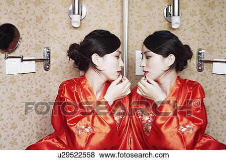 Stock Photo - Young woman looking in the mirror applying lipstick. Fotosearch