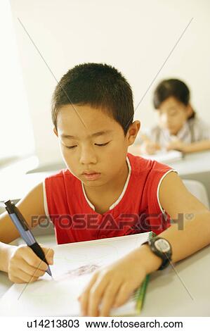 Stock Image - Close-up of a boy writing in a spiral notebook in the classroom. Fotosearch