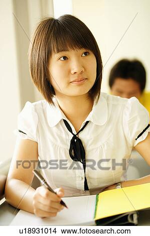 Picture - Close-up of a young woman writing on a notepad in a classroom. Fotosearch