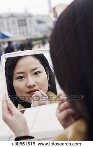 Stock Photograph - Close-up of a young woman looking into a mirror applying lipstick. Fotosearch