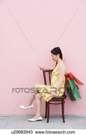 Stock Image - Side profile of a young woman sitting on a chair and using a mobile phone. Fotosearch