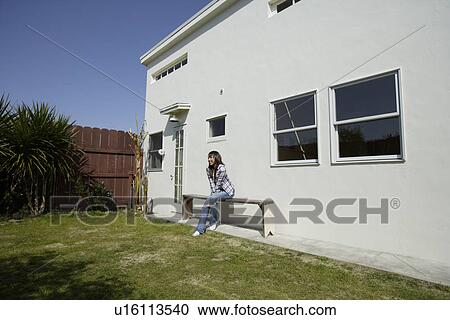 Stock Image - Young woman sitting on bench beside house. Fotosearch